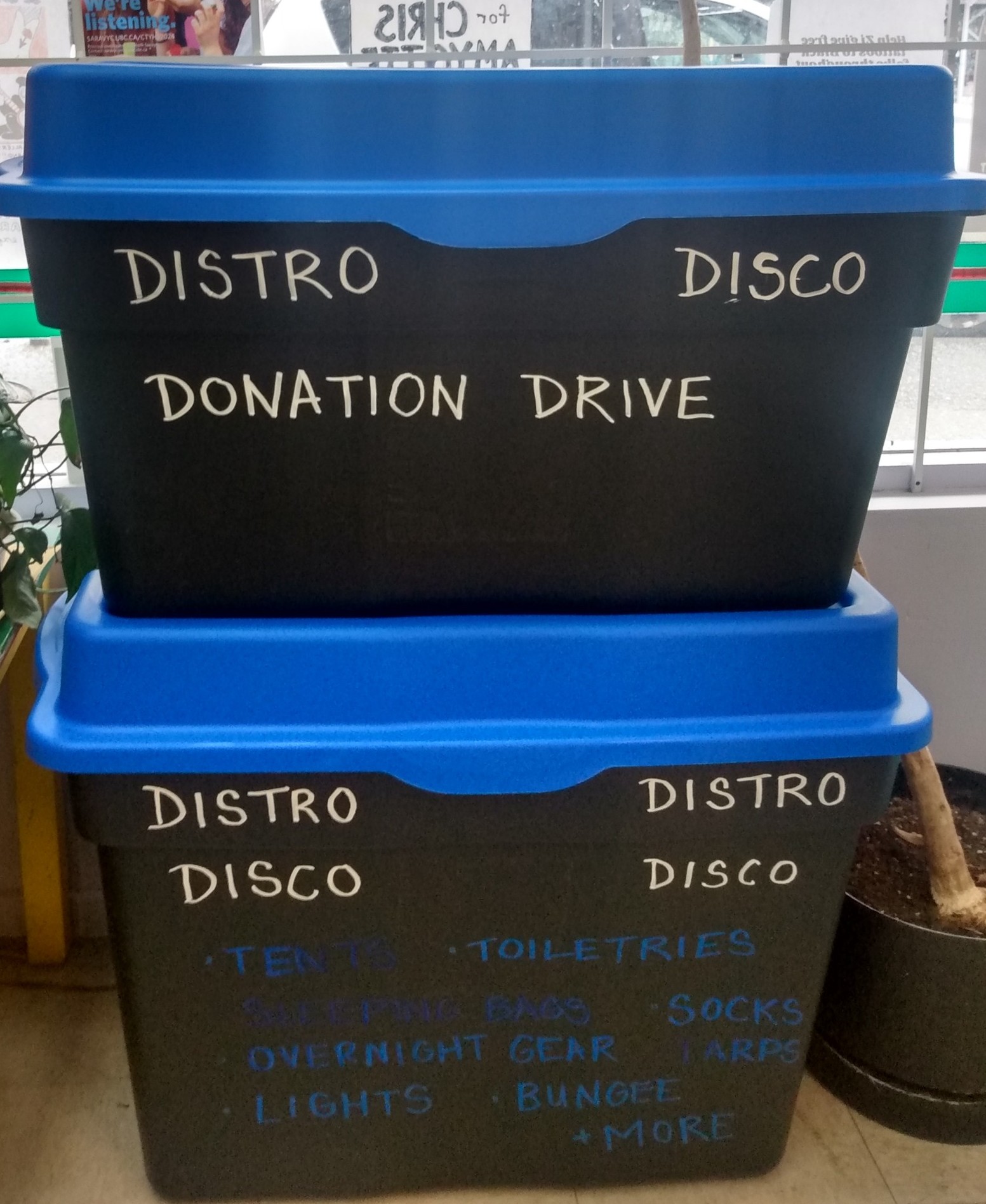 Two stacked black plastic bins with blue lids. Each has the words "DISTRO DISCO DONATION DRIVE" written in white on the front side. The lower bin has a bullet point list of needed donations written in dark blue, with some words smeared: "TENTS, TOILETRIES, SLEEPING BAGS, SOCKS, OVERNIGHT GEAR, TARPS, LIGHTS, BUNGEE + MORE"
